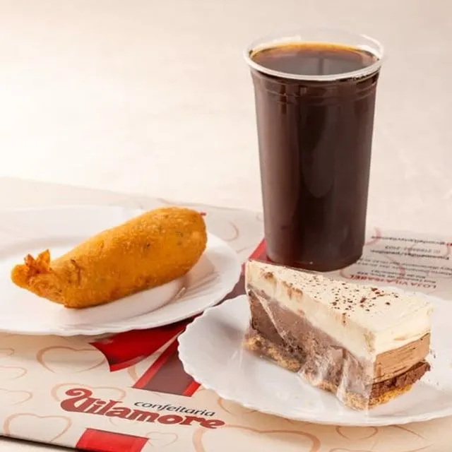 Salgado frito, copo de bebida gelada e uma fatia de bolo em camadas sobre papel com a marca Vilamore.