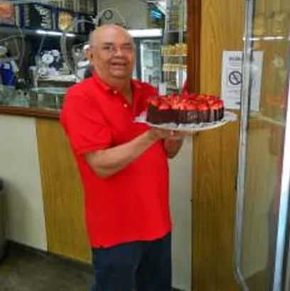 Seu Pedro, fundador da Vilamore, de camisa vermelha, segurando uma torta coberta de morangos no balcão da loja.